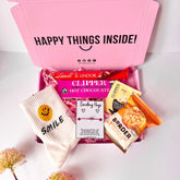 A pink gift box containing various items including a clippers hot chocolate sachet, a borders biscuit pack, a pair of smiley face crew socks, a sending hugs wish bracelet, a Lindor chocolate and a herbal tea bag.